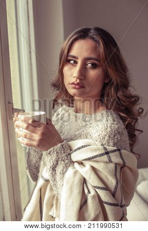Young Beautiful Woman Stands Near A Window, Wrapped In A Blanket And Drink Hot Coffee. Cozy. Autumn.