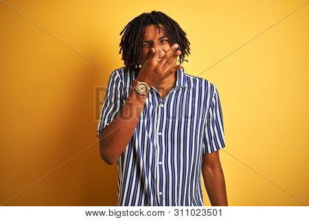 Afro man with dreadlocks wearing casual striped t-shirt over isolated yellow background smelling something stinky and disgusting, intolerable smell, holding breath with fingers on nose. Bad smells 