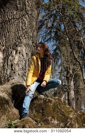 Outdoos Portrait Of Young Girl In Yellow Jacket Sitting Near Big Old Tree In The Park During Sunny D