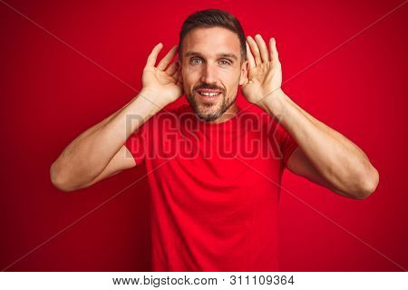 Young handsome man wearing casual t-shirt over red isolated background Trying to hear both hands on ear gesture, curious for gossip. Hearing problem, deaf
