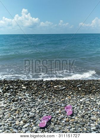 couple purple rubber flip-flops lie on the beach in the direction of the sea, rubber flip flops with striped sole lie on the beach, pebbly beach, turquoise expanse of water, the white foam of the surf, the straight line of the horizon, the azure sky, a se