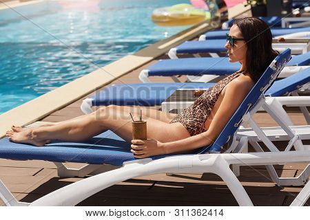 Slender Black Haired European Woman Lying On Blue Chaise Lounge At Resort, Wearing Trendy Sunglasses
