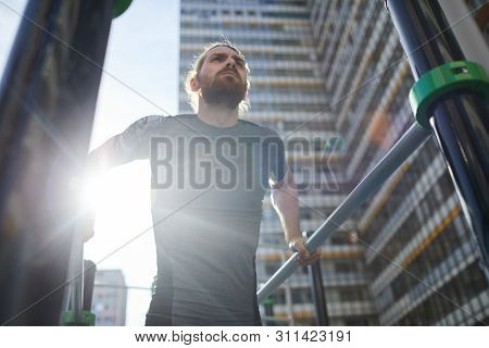 Below View Of Determined Bearded Athlete In Tshirt Hanging On Parallel Bars And Looking Into Distanc