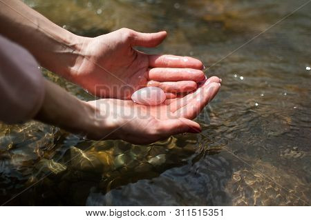 Woman Holding In Hand A Yoni Egg. Rose Quartz Crystal Egg On River Background. Female Health Concept