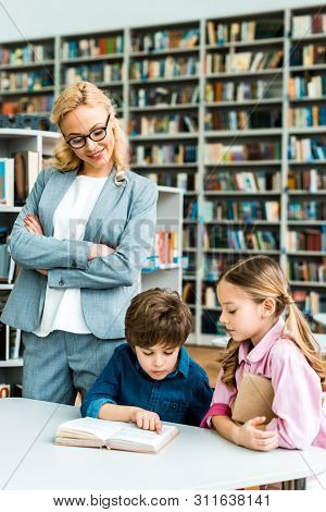 Cheerful Teacher In Glasses Standing With Crossed Arms And Looking At Cute Kids In Library