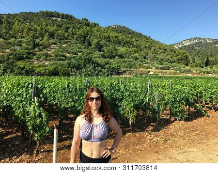 A Happy Young Woman Standing In Front Of A Sprawling Wine Vineyard Growing The Local Grk Grapes On K