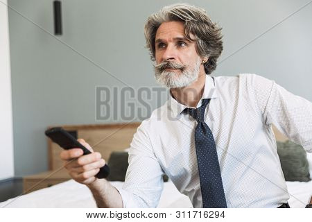 Image closeup of caucasian adult man wearing suit and tie holding remote control while watching television on bed in hotel apartment