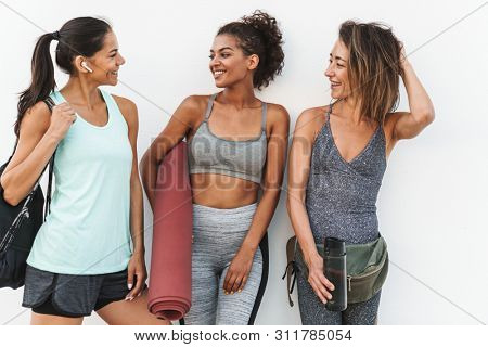 Photo of fitness multiethnic sportswomen in tracksuits smiling and standing over white wall outdoors