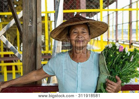 Maing Thauk, Myanmar - April 2019: Portrait Of An Old Burmese Woman In A Bamboo Hat And Flowers