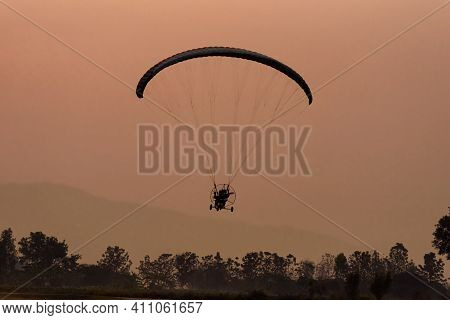 Silhouette Of The Paramotor Gliding And Flying In The Air Through Soft Sunlight Sky.