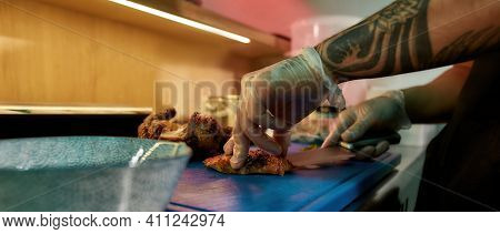 Chef With Tattoos Is Preparing Delicious Food. Cook Cuts Chicken On The Table Of An Asian Restaurant