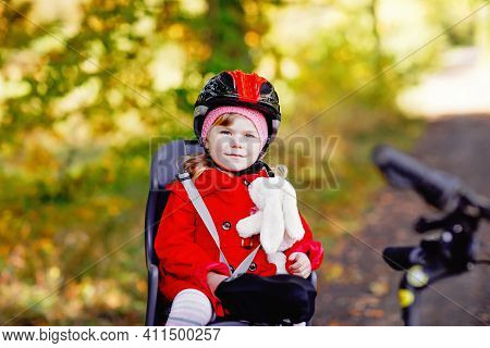 Little Toddler Girl With Security Helmet On Head Sitting In Bike Seat Of Her Mother Or Father Bicycl