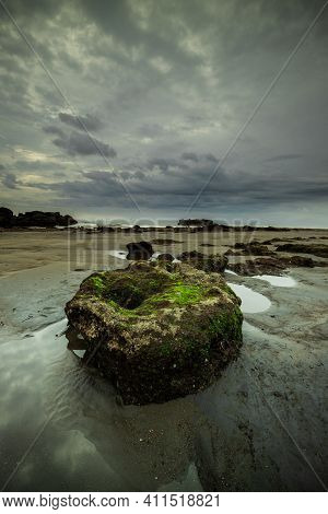 Landscape With Grey Cloudy Sky. Big Stone On Black Sand Beach. Stone Covered By Green Moss. Low Tide