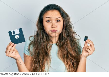 Young hispanic girl holding floppy disk and sdxc card puffing cheeks with funny face. mouth inflated with air, catching air. 