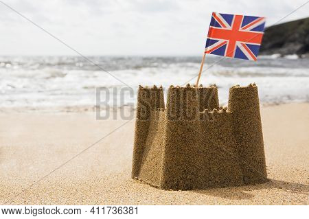 Sandcastle On Empty British Beach With Uk Union Jack Flag Flying