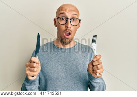Bald man with beard holding fork and knife ready to eat afraid and shocked with surprise and amazed expression, fear and excited face. 