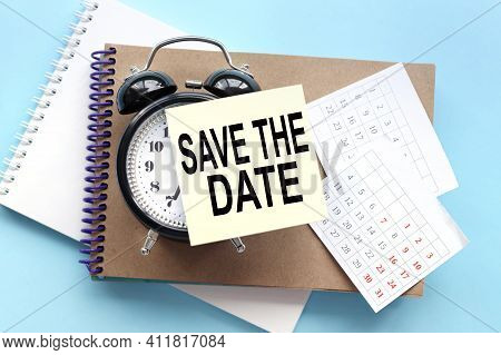 Save The Date. Text On The Sticker Attached To The Alarm Clock. Alarm Clock On Notepad On Blue Backg