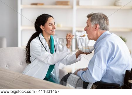 Medical Assistance For Elderly People With Disabilities. Young Nurse Feeding Senior Handicapped Man 