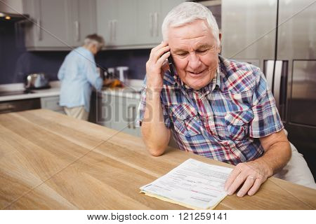 Senior man talking on phone and woman working in kitchen at home