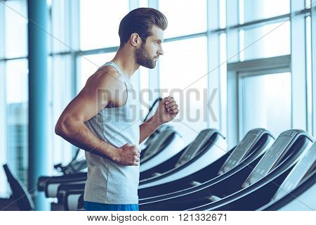 Running fast. Side view of young man in sportswear running on treadmill at gym