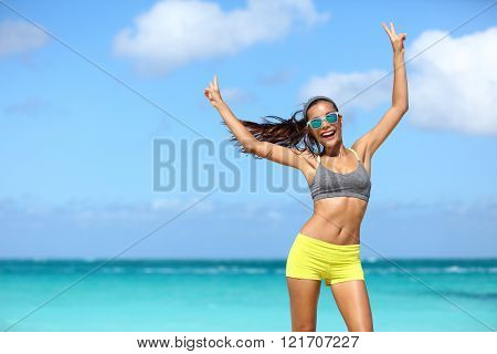 Happy cheerful fitness woman in sunglasses winning arms up doing v hand signs. Fit girl with slim body on sunny beach with raised arm showing peace sign in happiness of weight loss after workout. 