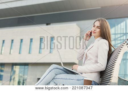 Businesswoman working on laptop. Caucasian insuranse agent checking email on computer, talking on mobile, sitting on the bench outdoors near modern office center