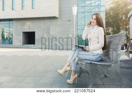 Smiling businesswoman talking on mobile and working with tablet. Caucasian insuranse agent checking email and consulting on smartphone, sitting on the bench outdoors in urban area