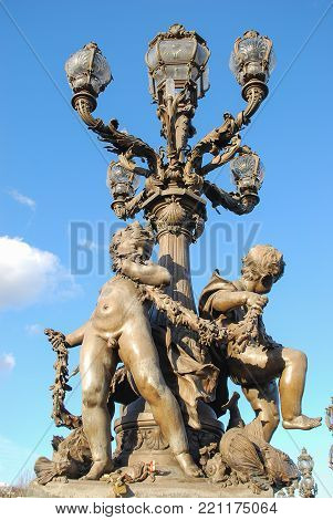 Boys nymph staue decorated under lamp or light on bridge in action standing and holds things in hand ,some fishes statue near feet with blackground blue sky and clouds.