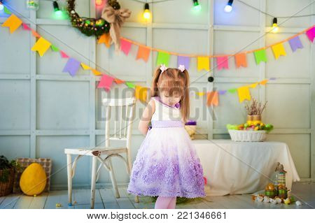 A little girl is standing in a decorated room, and downtrodden or punished her head, looking at the floor. Easter photo with a garland, eggs, a hare, a child.