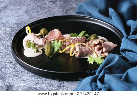 Vitello tonnato italian dish. Thin sliced veal with tuna sauce, capers and coriander served on black plate with blue textile over blue texture background. Close up