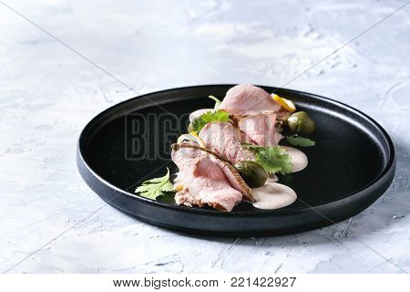 Vitello tonnato italian dish. Thin sliced veal with tuna sauce, capers and coriander served on black plate over gray texture background.