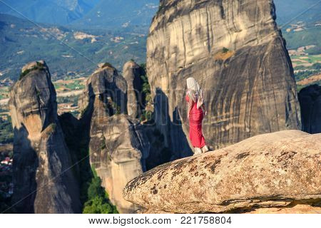 Alone girl in a red dress and scarf on the edge of the rock in Meteora. Creece in Thessaly. Pilgrimage in Greece