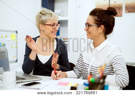 Close up of two smiling stylish business middle aged women working and having a conversation while sitting in the office one next to another.