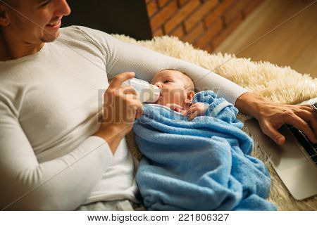 Man with baby. Young man feeding little baby while lying on bed and working on laptop