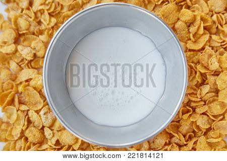 Cornflakes scattered on a wooden table. Round frame with metal bowl with milk lined with corn flakes.