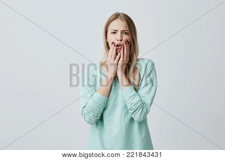 Shocked stunned blonde european woman with opened mouth, holding hands on cheeks, shocked by news she heard. Frightened terrified beautiful female model. Negative emotions