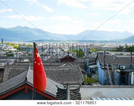 The Flag Of China Or Five-starred Red Flag With Rooftops Of The Old Town Of Lijiang In The Backgroun