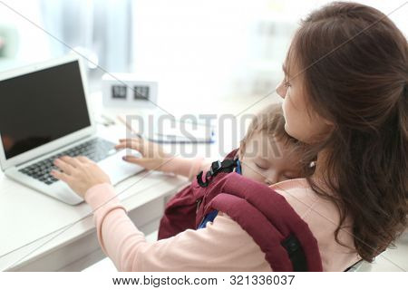 Young woman with baby in ergo carrier using laptop indoors