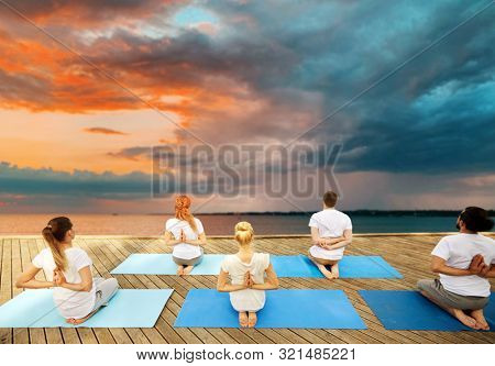 fitness, sport, yoga and healthy lifestyle concept - group of people exercising in reverse prayer pose on sea pier over sunset background