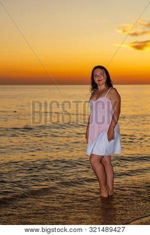 A Young Woman In A White Dress Stands On The Seashore At Sunset.