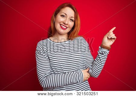 Young redhead woman wearing strapes navy shirt standing over red isolated background with a big smile on face, pointing with hand and finger to the side looking at the camera.