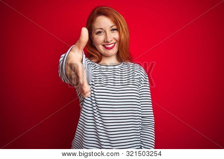 Young redhead woman wearing strapes navy shirt standing over red isolated background smiling friendly offering handshake as greeting and welcoming. Successful business.