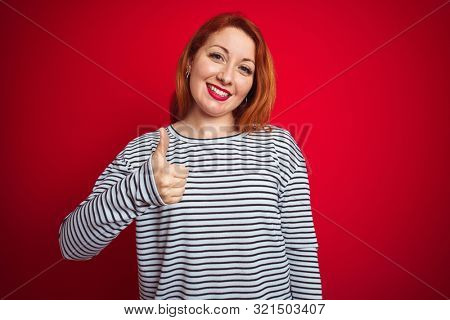Young redhead woman wearing strapes navy shirt standing over red isolated background doing happy thumbs up gesture with hand. Approving expression looking at the camera showing success.