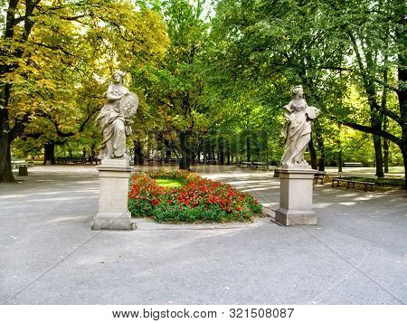 Two Sculptures Of Ancient Roman Goddesses On A Background Of Yellow-orange-green Trees And Red Flowe