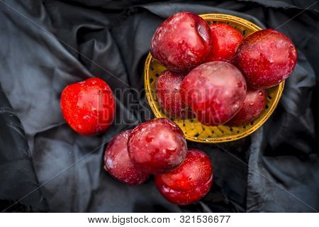 Raw Organic Red Ripe Plums In A Brown-colored Basket On Black Fabric On The Wooden Surface.