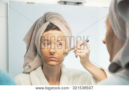 Young Woman Puts Clay Mask On Her Face With Brush Looking In Mirror In Bathroom. The Concept Of Self
