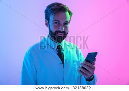 Portrait of a smiling young man doctor wearing uniform standing isolated over pink-blue haze backgroundm using mobile phone