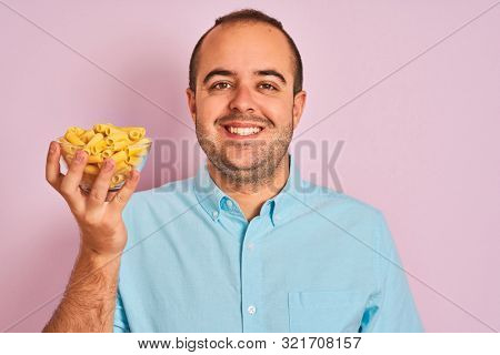Young man holding bowl with macaroni pasta standing over isolated pink background with a happy face standing and smiling with a confident smile showing teeth
