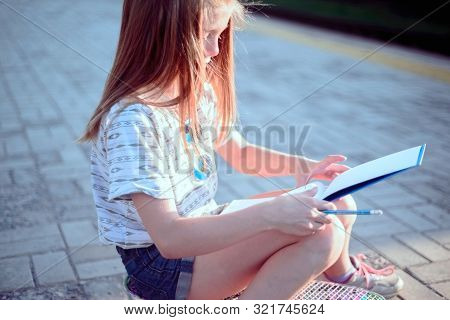 Beautiful Little Girl Sitting On A Skateboard And Writing In A Tetrad. Outdoor Lifestyle