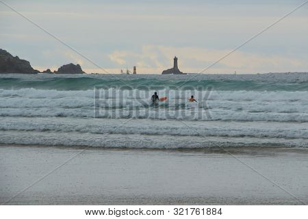 Two Surfers At Baie Des Trepasses Beach In Plogoff With La Vieille Lighthouse In The Background